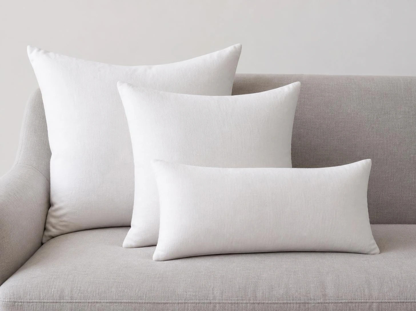 Three plain white pillows layered on a light grey sofa: a large square at the back, a smaller square in front and a lumbar in the middle