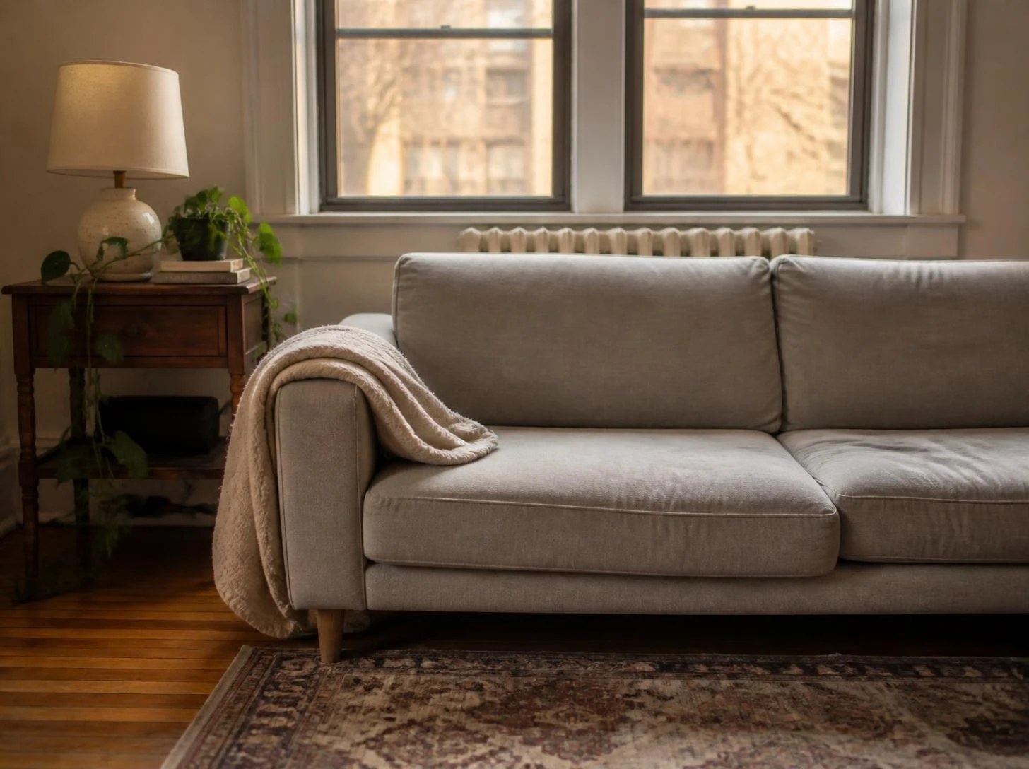 A bare grey sofa in a prewar apartment, with a throw over one arm, a lamp on a wooden side table and a radiator under the window